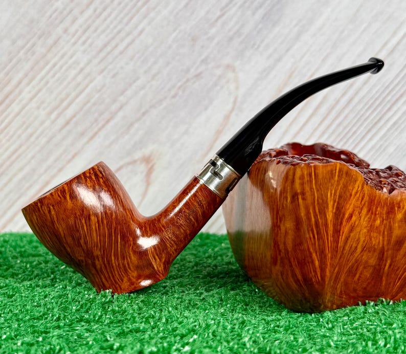 May include: A polished brown wooden tobacco pipe with a smooth, curved grain pattern, featuring a black acrylic stem and a silver-toned metal band connecting the bowl to the stem, resting against a large, textured block of matching brown wood.