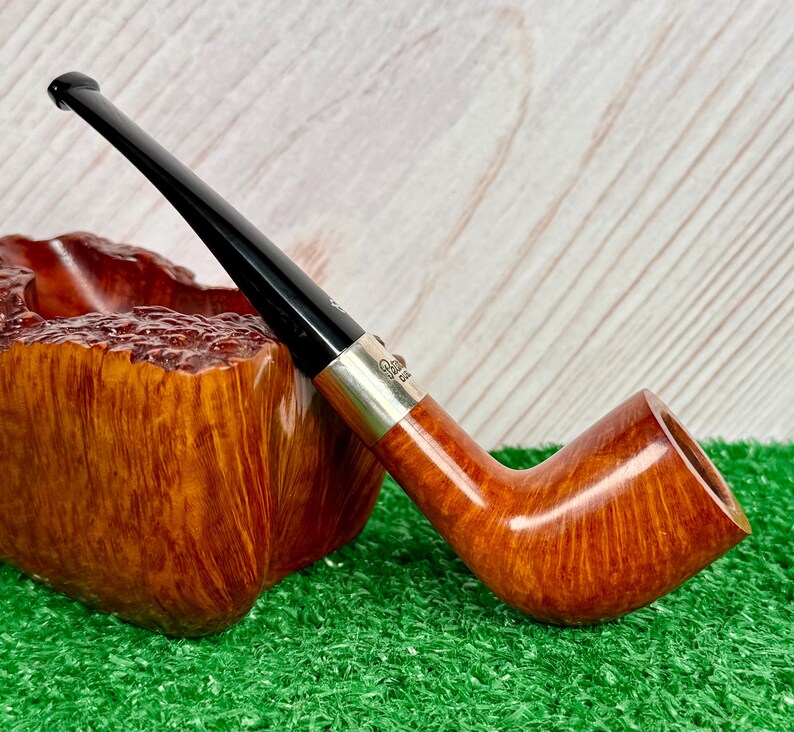 Puede incluir: Una pipa de tabaco con un hornillo de madera de color marr&oacute;n claro liso y una boquilla negra larga y delgada. Una banda de metal plateado conecta el hornillo con la boquilla, con el texto grabado Peterson Dublin. La pipa est&aacute; en diagonal.
