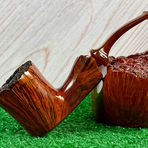 May include: A handcrafted, wooden tobacco pipe with a rich, reddish-brown finish. The pipe features a square bowl with a textured rim and a curved stem. The bowl is resting on a textured wooden base, and the pipe is set against a green surface.