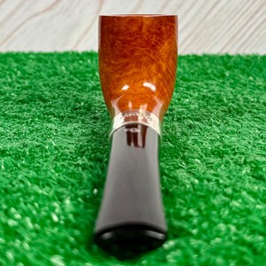 Puede incluir: Una pipa de tabaco con un hornillo de madera de color marr&oacute;n claro pulido con veta visible, conectada a una boquilla de color marr&oacute;n oscuro mediante una banda de metal plateado. La pipa est&aacute; colocada verticalmente con la boquilla abajo.