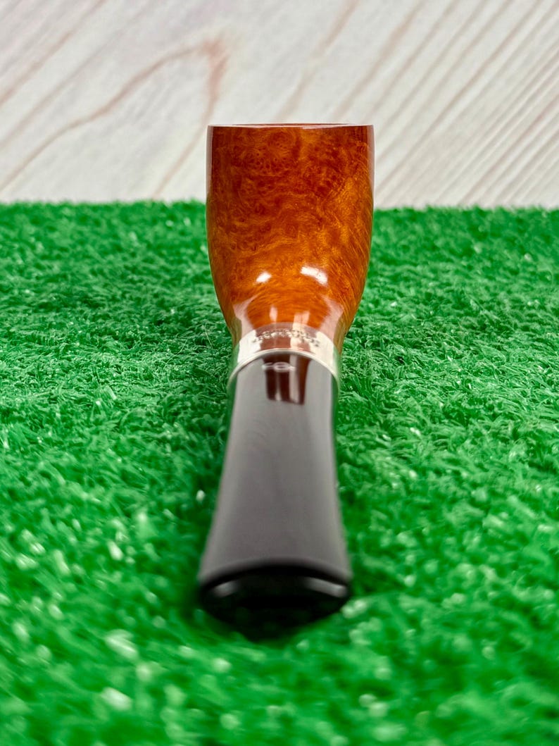 Puede incluir: Una pipa de tabaco con un hornillo de madera de color marr&oacute;n claro pulido con veta visible, conectada a una boquilla de color marr&oacute;n oscuro mediante una banda de metal plateado. La pipa est&aacute; colocada verticalmente con la boquilla abajo.