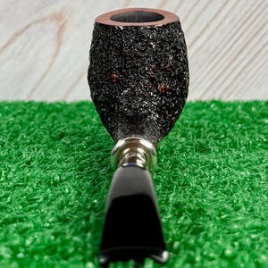 May include: A close-up of a tobacco pipe with a dark, textured bowl and a black stem. The bowl has a rough, bark-like appearance, and the stem is smooth and glossy. The pipe is set against a green background.