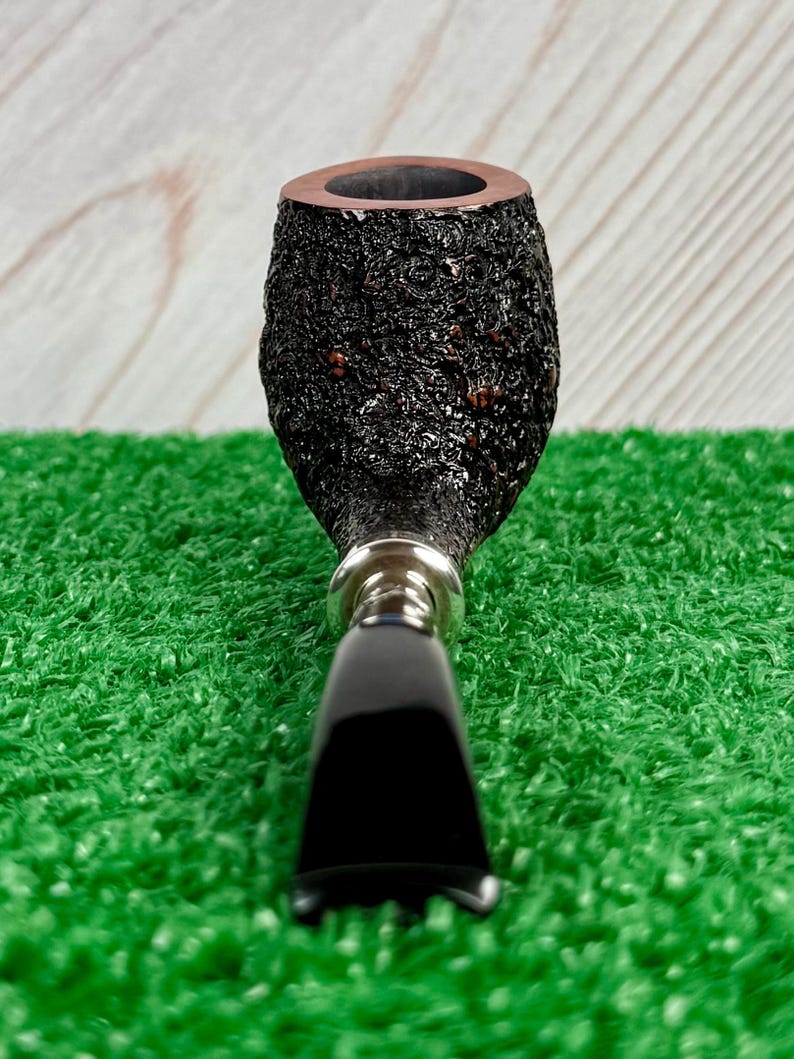May include: A close-up of a tobacco pipe with a dark, textured bowl and a black stem. The bowl has a rough, bark-like appearance, and the stem is smooth and glossy. The pipe is set against a green background.