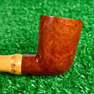May include: A handcrafted tobacco pipe with a curved, reddish-brown bowl and a light-colored stem. The bowl features a unique wood grain pattern. The stem is connected to a light-colored bamboo-like section. The pipe rests on a green surface.