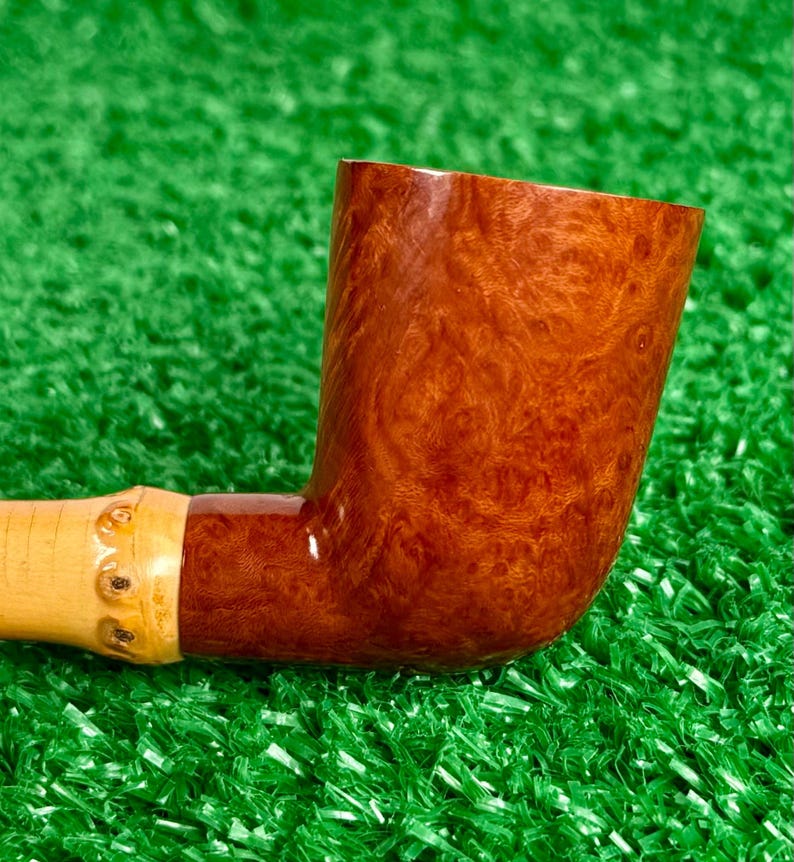 May include: A handcrafted tobacco pipe with a curved, reddish-brown bowl and a light-colored stem. The bowl features a unique wood grain pattern. The stem is connected to a light-colored bamboo-like section. The pipe rests on a green surface.