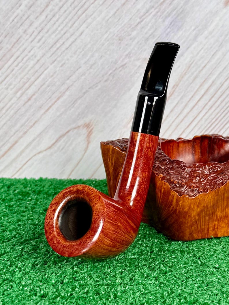 May include: A polished tobacco pipe with a smooth, reddish-brown wooden bowl and a sleek black acrylic stem. The pipe is positioned next to a carved, textured wooden block that features a natural, rugged grain and a deep, hollowed-out center.