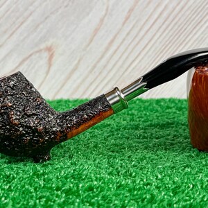 May include: A tobacco pipe with a dark, textured bowl and a smooth, curved stem. The stem is black with a silver band near the bowl. The pipe rests on a green surface, with a wooden block in the background.