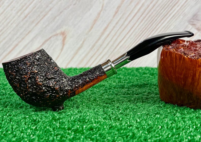 May include: A tobacco pipe with a dark, textured bowl and a smooth, curved stem. The stem is black with a silver band near the bowl. The pipe rests on a green surface, with a wooden block in the background.