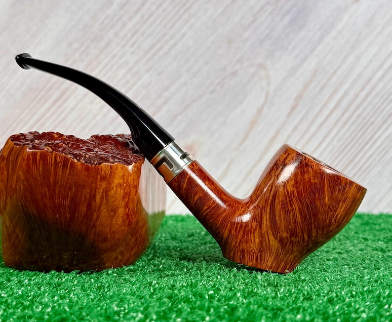 May include: A tobacco pipe with a polished brown wooden bowl featuring a visible grain pattern, a curved black stem, and a silver-colored metal band connecting the two sections. The pipe is positioned next to a rough, textured wooden block.