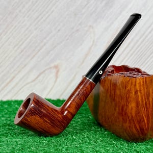 May include: A brown wooden tobacco pipe with a smooth, polished finish and a long, slender black stem. The pipe features a silver-colored band at the junction of the bowl and stem, with the text "RAY WOODS" and "FLAME GRAIN" engraved on the side.