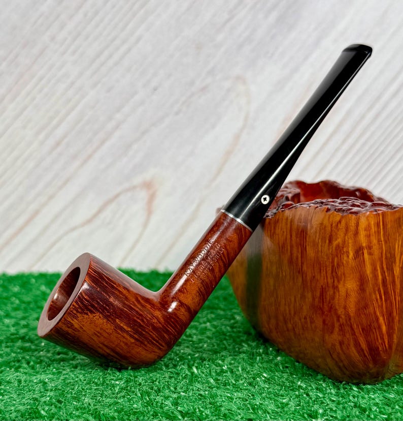 May include: A brown wooden tobacco pipe with a smooth, polished finish and a long, slender black stem. The pipe features a silver-colored band at the junction of the bowl and stem, with the text "RAY WOODS" and "FLAME GRAIN" engraved on the side.