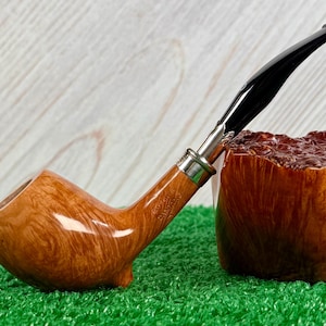 May include: A wooden tobacco pipe with a curved stem and a black mouthpiece. The pipe has a light brown finish with a silver band near the mouthpiece. The bowl is a darker brown color and has a textured surface. The pipe is resting on a green surface.