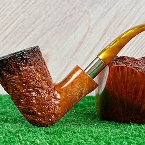 May include: A brown tobacco pipe with a textured bowl and a curved, amber-colored stem. The pipe has a silver band and is resting on a green surface. The bowl is dark brown at the top.