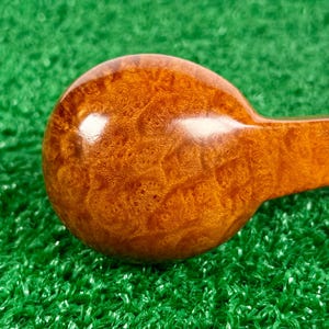 May include: A polished, round wooden object with a rich, golden-brown burl grain pattern. The surface is smooth and glossy, showing intricate swirling wood textures. A portion of a straight handle extends from the side of the circular head.