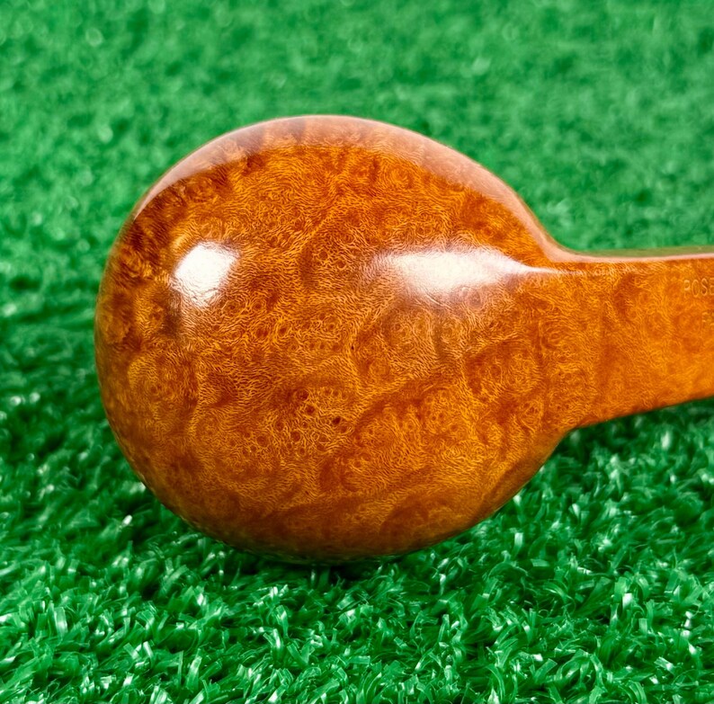 May include: A polished, round wooden object with a rich, golden-brown burl grain pattern. The surface is smooth and glossy, showing intricate swirling wood textures. A portion of a straight handle extends from the side of the circular head.