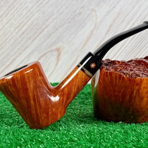 Pode incluir: Um cachimbo de tabaco de madeira com um bocal preto curvo e uma tigela castanha. O cachimbo tem um acabamento polido, mostrando o grão da madeira. O cachimbo está sobre uma superfície verde.