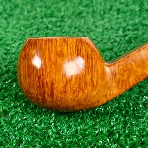 May include: A polished tobacco pipe crafted from light brown wood with a visible natural grain pattern. The bowl is rounded and smooth, featuring a glossy finish that reflects light, connected to a tapered stem extending toward the right side.