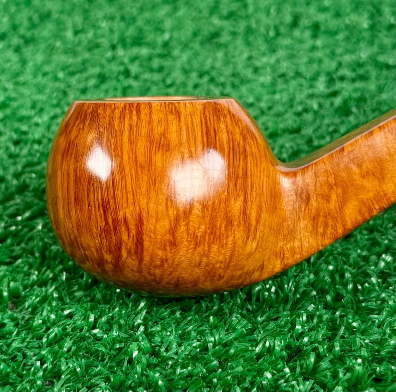 May include: A polished tobacco pipe crafted from light brown wood with a visible natural grain pattern. The bowl is rounded and smooth, featuring a glossy finish that reflects light, connected to a tapered stem extending toward the right side.