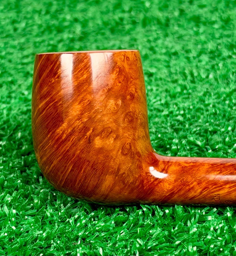 Op de afbeelding: Een gepolijste tabakspijp gemaakt van lichtbruin hout met een opvallend, wervelend nerfpatroon. De kop is cilindrisch en glad en gaat over in een horizontale steel. Het hout vertoont natuurlijke knoesten en amberkleurige accenten.