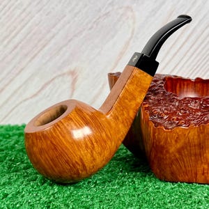 May include: A polished light brown wooden tobacco pipe with a smooth, rounded bowl and a long, curved black stem. The stem features a small gold-colored logo detail near the junction with the wooden shank, showcasing a natural wood grain finish.