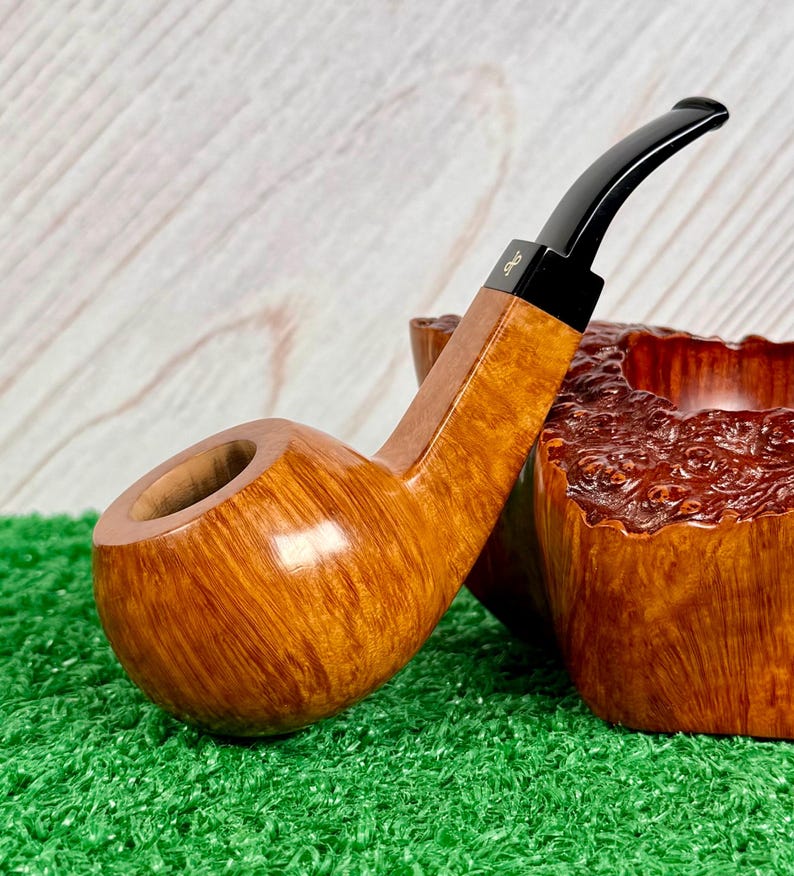 May include: A polished light brown wooden tobacco pipe with a smooth, rounded bowl and a long, curved black stem. The stem features a small gold-colored logo detail near the junction with the wooden shank, showcasing a natural wood grain finish.
