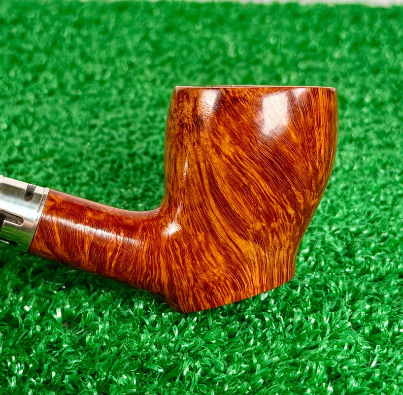 May include: A polished brown tobacco pipe with a prominent wood grain pattern and a smooth, glossy finish. The pipe features a cylindrical bowl and a slender stem connected by a silver-colored metal band, showcasing natural wood textures.