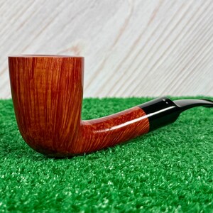 May include: A tobacco pipe featuring a polished, light brown wooden bowl with a visible vertical grain pattern. The pipe has a smooth, curved shank that transitions into a sleek, black acrylic stem with a tapered mouthpiece for smoking.