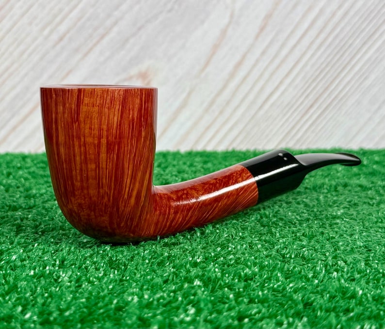 May include: A tobacco pipe featuring a polished, light brown wooden bowl with a visible vertical grain pattern. The pipe has a smooth, curved shank that transitions into a sleek, black acrylic stem with a tapered mouthpiece for smoking.