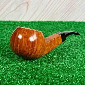 May include: A tobacco pipe featuring a polished, light brown wooden bowl with a visible grain pattern. The pipe has a square-shaped wooden shank connected to a smooth, black acrylic stem. The design includes a small, metallic logo on the stem.