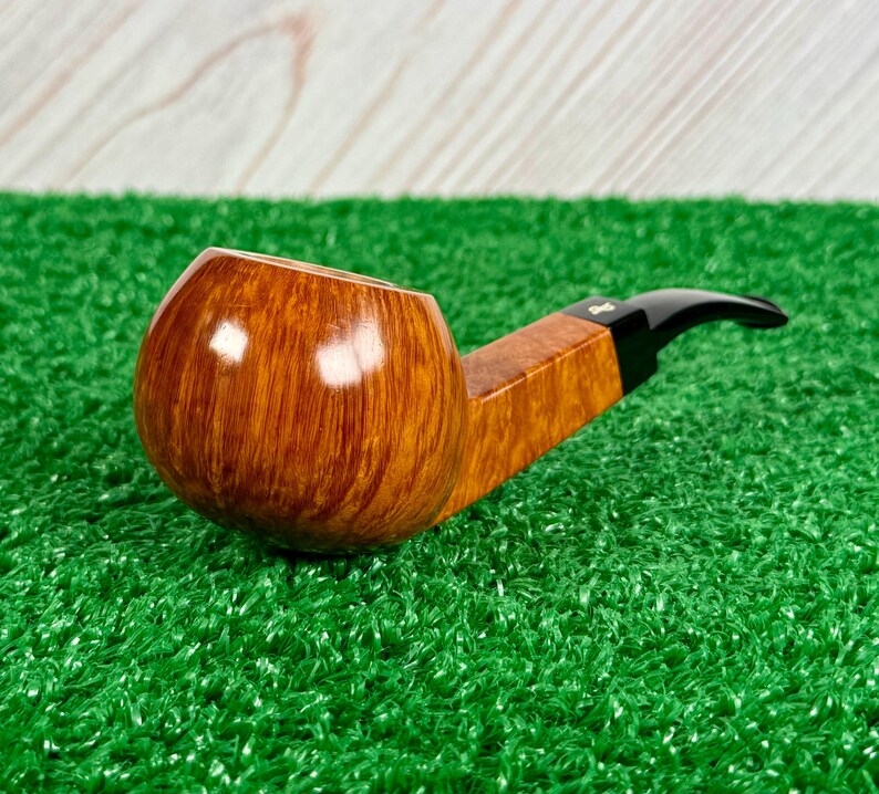 May include: A tobacco pipe featuring a polished, light brown wooden bowl with a visible grain pattern. The pipe has a square-shaped wooden shank connected to a smooth, black acrylic stem. The design includes a small, metallic logo on the stem.