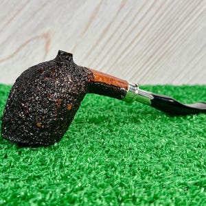 May include: A dark brown, textured tobacco pipe with a curved stem. The pipe features a wooden bowl and a black mouthpiece. The pipe rests on a green surface, with a light wood background.