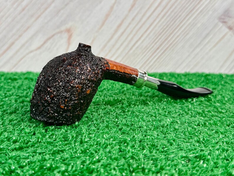 May include: A dark brown, textured tobacco pipe with a curved stem. The pipe features a wooden bowl and a black mouthpiece. The pipe rests on a green surface, with a light wood background.