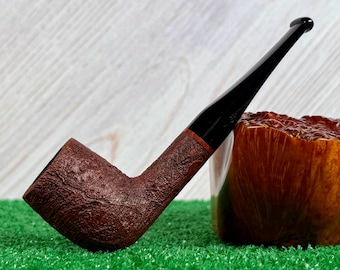 May include: A tobacco pipe with a dark brown, textured, rusticated bowl and a smooth, glossy black stem. The pipe is positioned at an angle, showcasing the contrast between the rough, porous surface of the bowl and the sleek, polished finish of the stem.