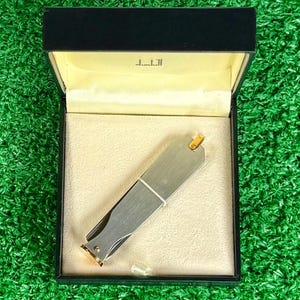 Pode incluir: Um cortador de unhas prateado com detalhes dourados é exibido dentro de uma caixa preta com interior creme. A caixa tem a marca "Dunhill" impressa no interior da tampa. O cortador de unhas é preso com uma faixa branca.