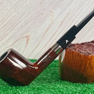 Pode incluir: Um cachimbo Ascort castanho com boquilha preta e faixa prateada. O cachimbo está sobre uma superfície verde com um bloco de madeira ao fundo. O texto "Ascort" é visível no cachimbo.