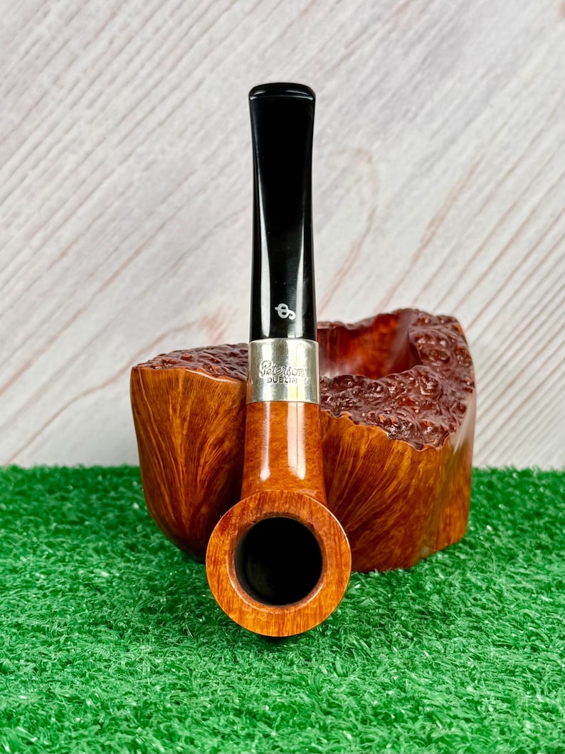 Puede incluir: Una pipa de tabaco con un hornillo de madera de color marr&oacute;n claro liso y una boquilla negra. Una banda de plata conecta la boquilla con el hornillo, con el texto grabado Peterson Dublin. Un peque&ntilde;o logotipo blanco es visible en la boquilla.