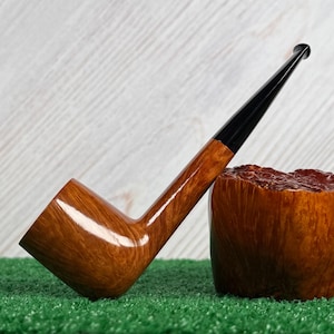 May include: A tobacco pipe with a polished light brown wooden bowl and a long, slender black stem. The pipe is positioned next to a large, textured, dark brown wooden block that shows a natural, rugged grain pattern on its top surface.