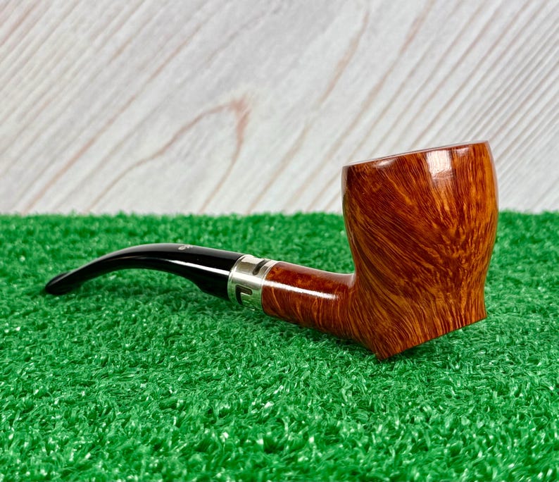 May include: A tobacco pipe featuring a polished brown wood grain bowl with a smooth finish. The stem is made of black acrylic and connects to the bowl via a silver-colored metal band. The pipe has a curved shape and a wide, rounded bowl top.