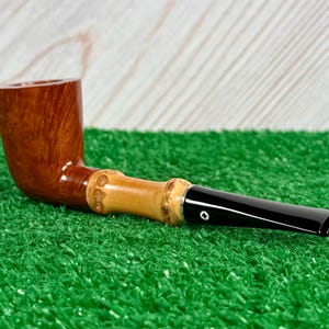 May include: A wooden tobacco pipe with a curved, brown bowl and a light-colored bamboo stem. The stem has carved details and connects to a black mouthpiece. The pipe rests on a green surface.