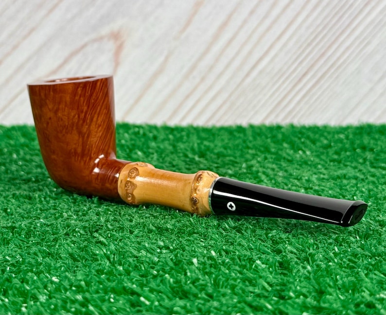 May include: A wooden tobacco pipe with a curved, brown bowl and a light-colored bamboo stem. The stem has carved details and connects to a black mouthpiece. The pipe rests on a green surface.