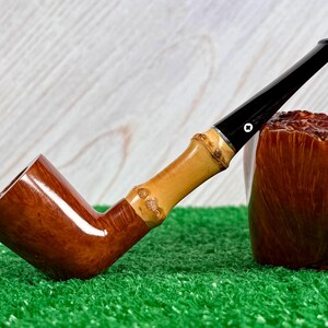 May include: A wooden tobacco pipe with a bamboo stem and a black mouthpiece. The pipe has a curved design and rests on a green surface. A brown, textured object is next to the pipe.