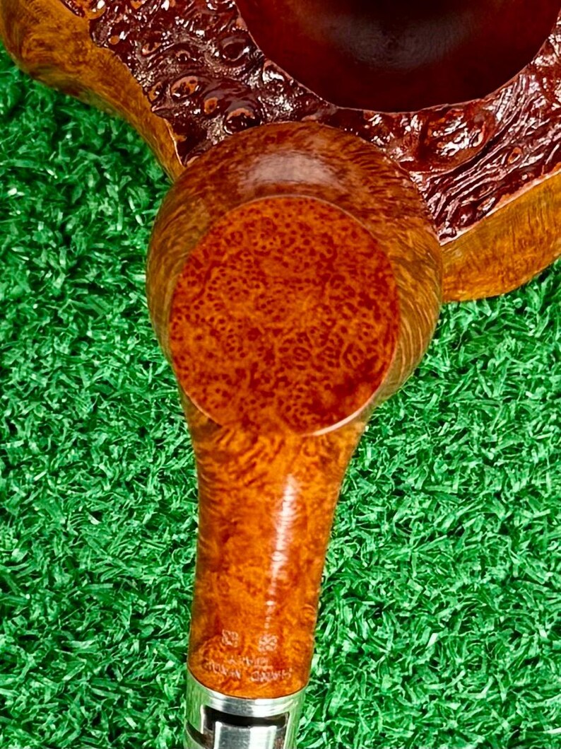 May include: A tobacco pipe featuring a polished, light brown briar wood bowl with a distinct burl grain pattern. The stem is connected to a silver-colored metal band, and the wood surface displays a smooth, glossy finish with visible natural textures.