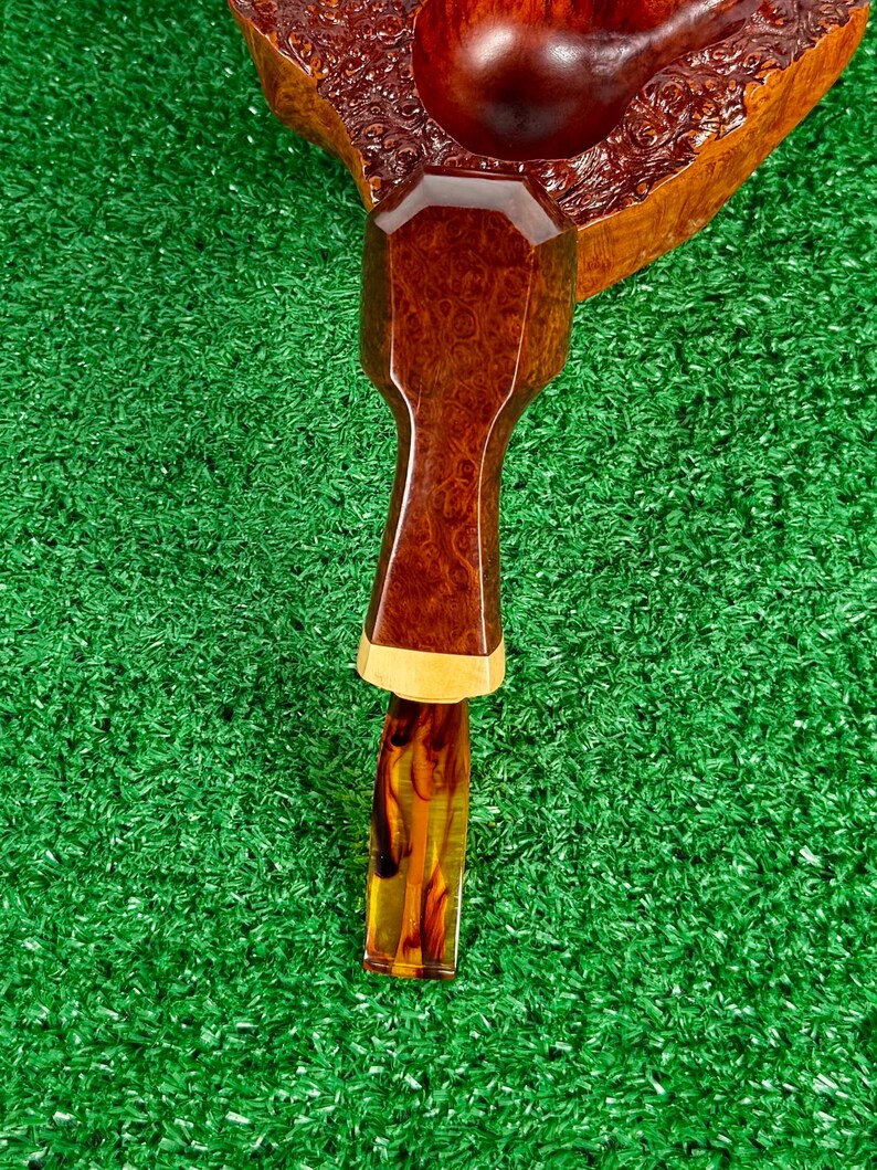 Pode incluir: Um cachimbo de tabaco com fornilho de madeira castanho-escura polida com um padr&atilde;o de gr&atilde;o de madeira texturizado. A haste &eacute; composta por uma sec&ccedil;&atilde;o de madeira castanha facetada ligada a um anel de madeira clara e uma boquilha de acr&iacute;lico &acirc;mbar transl&uacute;cido.