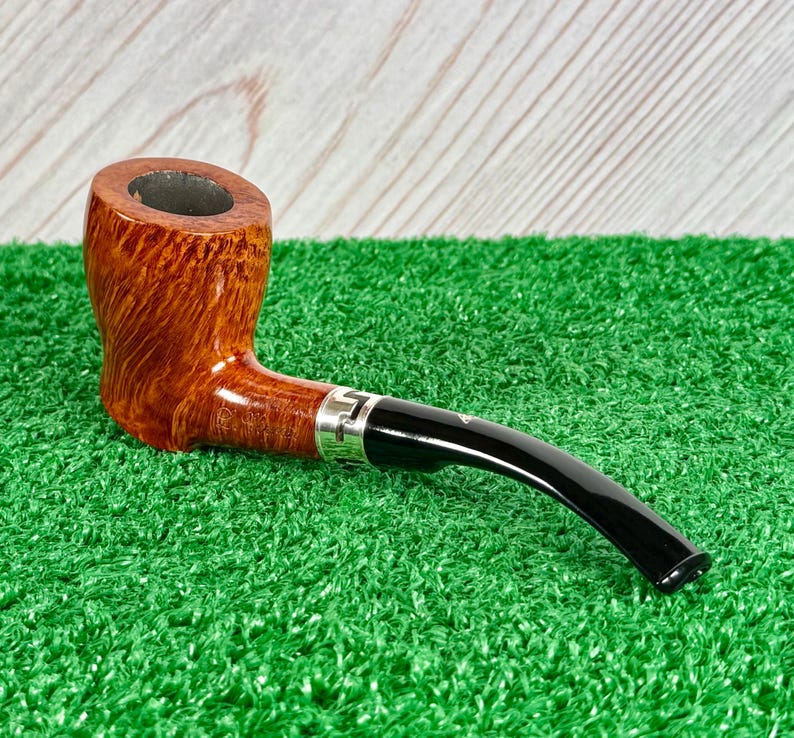 May include: A tobacco pipe with a polished brown wooden bowl featuring a visible grain pattern, a silver-colored metal band with an engraved geometric design, and a smooth, curved black acrylic stem extending from the side of the bowl.