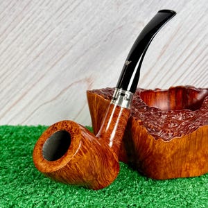May include: A tobacco pipe with a polished light brown wood grain bowl and a curved black stem featuring a silver-colored metal band. The pipe is positioned next to a matching, rough-textured, natural wood burl pipe rest with a deep central cavity.