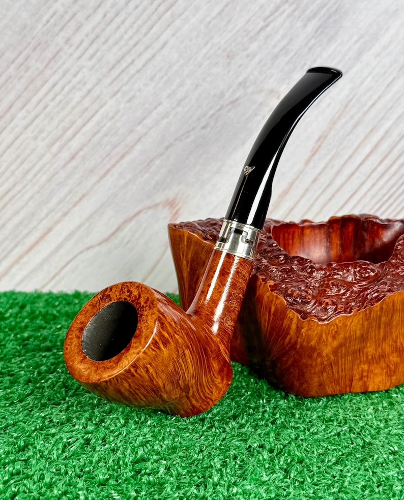 May include: A tobacco pipe with a polished light brown wood grain bowl and a curved black stem featuring a silver-colored metal band. The pipe is positioned next to a matching, rough-textured, natural wood burl pipe rest with a deep central cavity.