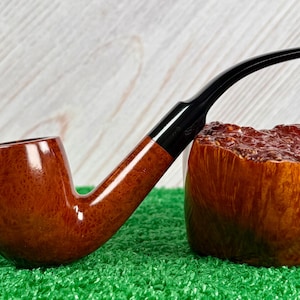Pode incluir: Um cachimbo de tabaco castanho com um bocal preto curvo. O cachimbo tem um acabamento brilhante e repousa sobre um leito de relva artificial verde. O cachimbo está ao lado de um bloco de madeira com uma superfície texturizada.