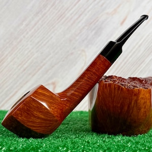 Pode incluir: Um cachimbo de tabaco de madeira castanha com boquilha preta, apoiado num bloco de madeira texturizado. O cachimbo e o bloco estão sobre uma superfície verde e um fundo de madeira branca.