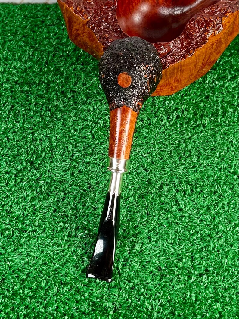 May include: A tobacco pipe with a dark, textured bowl and a wooden stem. The stem has a silver band and leads to a black, angled mouthpiece. The pipe rests on a wooden stand with a reddish-brown finish, set against a green background.