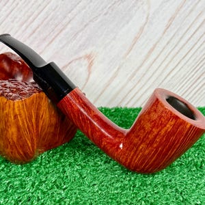 May include: A tobacco pipe with a polished reddish-brown wooden bowl and a smooth black acrylic stem. The pipe features a distinct grain pattern on the wood and is positioned next to a textured, burl-like wooden block with a deep amber finish.